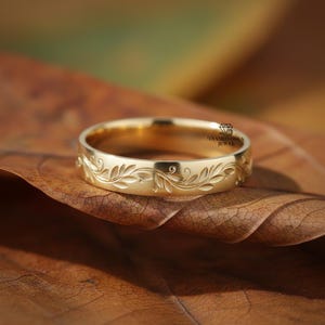 以下が含まれることがあります： 金色のリングで、葉のフローラルパターンが特徴です。リングは茶色の葉の上に置かれ、背景はぼやけています。リングは滑らかで磨かれた仕上げで、詳細な彫刻デザインが施されています。リングはジュエリーです。