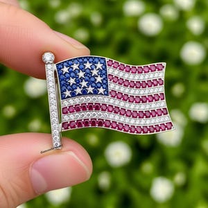 May include: A brooch in the shape of the American flag. The flag design features a blue field with white stars, red and white stripes, and a silver-colored pole. The brooch is adorned with gemstones.