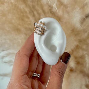 May include: Gold ear cuff with a row of faux pearls and rhinestones. The cuff is designed to wrap around the upper part of the ear.