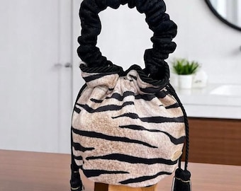 Puede incluir: Un bolso con cordón con un estampado de rayas de tigre en beige y negro. El bolso tiene un asa negra fruncida y un cordón negro con borlas. El bolso está sobre un pequeño soporte de madera.