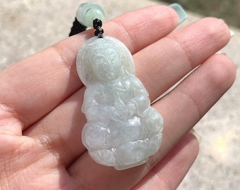 May include: A pale green jade pendant carved as a seated Buddha. The pendant hangs from a black cord with a green bead. The Buddha figure has detailed facial features. The pendant is held in a hand.