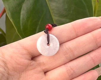 May include: A circular white jade pendant with a central hole, suspended from a black cord with a red bead. The jade has a translucent appearance. The pendant is a traditional design.