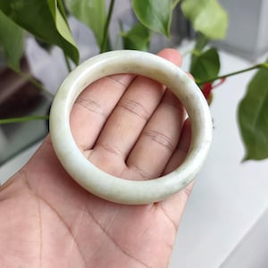 以下が含まれることがあります： 淡い緑色の翡翠バングル。円形のブレスレットは手で持たれており、自然な色のバリエーションを示しています。翡翠は半透明で、微妙な縞模様があります。