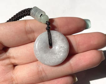 May include: A circular, pale grey jade pendant with a central hole, hanging from a black woven cord. The cord is attached to a clear, cylindrical bead and a small, green jade bead. The pendant has a smooth, polished finish.