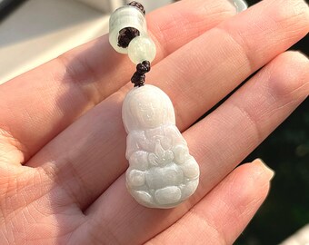 May include: A jade pendant with a carved Buddha figure. The pendant is mainly white with soft green tones, suspended from a dark brown cord. The Buddha figure has detailed facial features and folds in the carving.