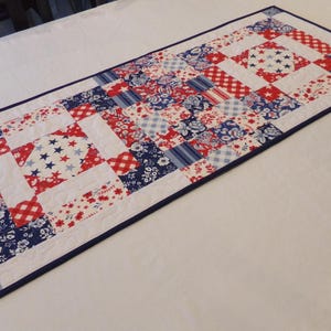 May include: A rectangular table runner with a patchwork design in red, white, and blue. The runner features squares with floral, star, and plaid patterns. A dark blue border frames the runner, which is likely made of fabric.