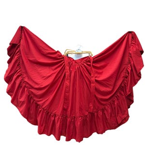 Pode incluir: Uma saia vermelha vibrante com uma bainha larga com babados e uma fita a condizer. A saia é exibida num cabide sobre um fundo branco, mostrando o seu design fluido e a sua forma completa. O tecido parece leve.