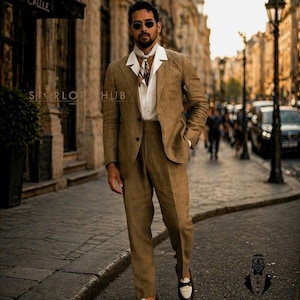 May include: A man wearing a light brown suit with a white shirt and patterned scarf, walking on a cobblestone street. The suit has a two-button jacket and matching trousers. He is wearing sunglasses and brown and white loafers. The image is taken in an urban setting.