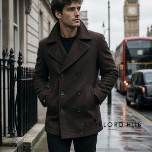 May include: A dark brown double-breasted coat with a wide collar and large buttons. The coat has two front pockets and a small logo on the lower right side. The coat is worn with black pants. The background shows a street scene with a red double-decker bus and Big Ben.