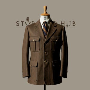 Può includere: Giacca marrone con quattro tasche frontali e bottoni dorati, esposta su un manichino. La giacca ha un design classico con una camicia con colletto e cravatta a righe sotto. Il logo "STYLE HUB" è visibile sullo sfondo.