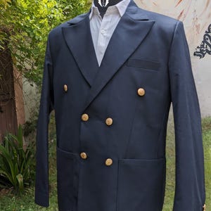 May include: A navy blue double-breasted blazer with gold-colored buttons. The blazer has a classic design with a notched lapel and two front pockets. It is displayed on a mannequin with a white shirt underneath. The background features greenery and a mural.