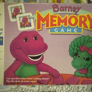 May include: A Barney Memory Game box featuring Barney the dinosaur and a green dinosaur character. The box has the text "Your First Memory!" and "For Ages 3 to 6". The game includes matching picture cards.