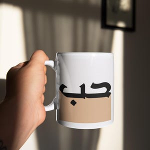 Puede incluir: Taza de cerámica blanca con un rectángulo marrón y la palabra árabe "حب" (que significa "amor") en letras negras.