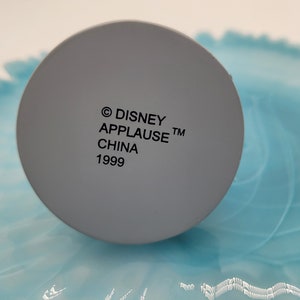 May include: A close-up of a grey, circular object with the text "&copy; DISNEY APPLAUSE&trade; CHINA 1999" printed in black. The object is set against a light blue and white background.