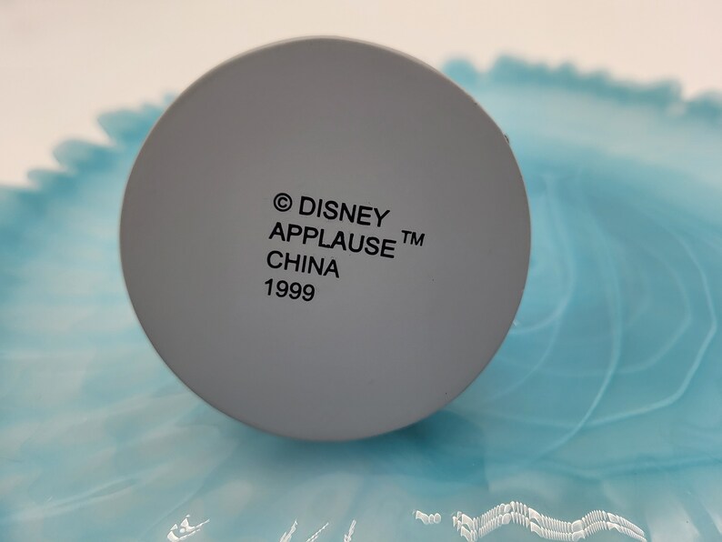 May include: A close-up of a grey, circular object with the text "&copy; DISNEY APPLAUSE&trade; CHINA 1999" printed in black. The object is set against a light blue and white background.