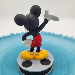 May include: A Mickey Mouse figurine in black, red, yellow, and white, stands on a grey film reel. The figure is posed with one arm raised, against a light blue background.