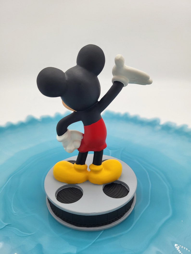 May include: A Mickey Mouse figurine in black, red, yellow, and white, stands on a grey film reel. The figure is posed with one arm raised, against a light blue background.