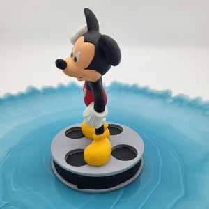 May include: A Mickey Mouse figurine, in his iconic outfit, stands on a grey film reel. Mickey is wearing yellow shoes, white gloves, red shorts, and a black body. The background is a light blue, swirled surface.
