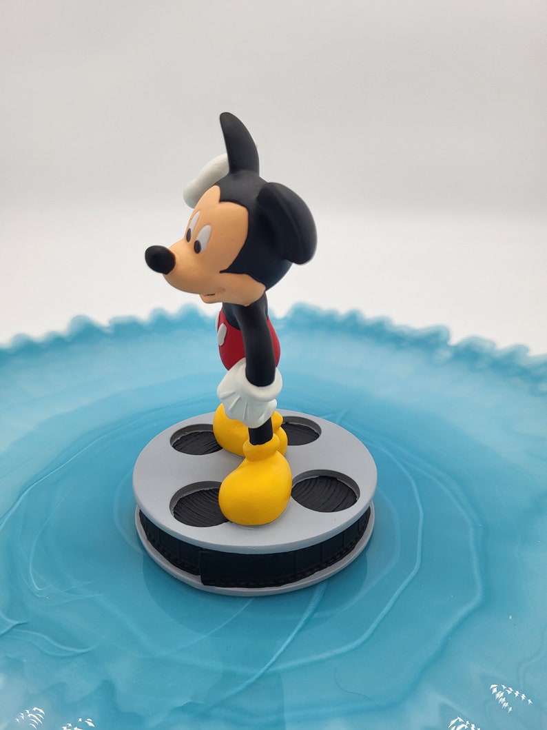 May include: A Mickey Mouse figurine, in his iconic outfit, stands on a grey film reel. Mickey is wearing yellow shoes, white gloves, red shorts, and a black body. The background is a light blue, swirled surface.