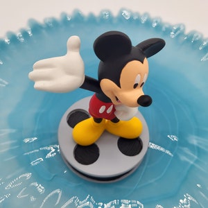 May include: A Mickey Mouse figurine, with black ears, red shorts, yellow shoes, and white gloves, stands on a grey film reel. The figurine is set against a light blue, ruffled background.