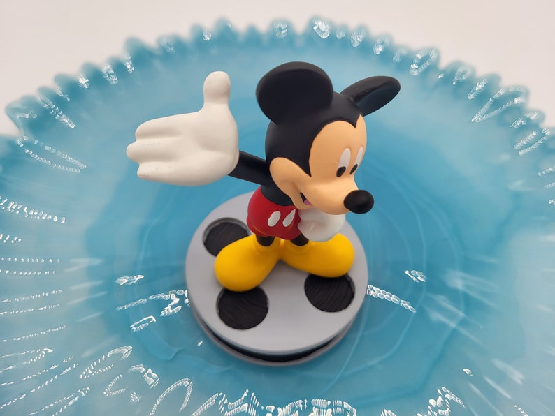 May include: A Mickey Mouse figurine, with black ears, red shorts, yellow shoes, and white gloves, stands on a grey film reel. The figurine is set against a light blue, ruffled background.