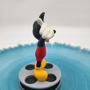 May include: A Mickey Mouse figurine, in his iconic outfit, stands on a grey film reel. Mickey is wearing red shorts, yellow shoes, white gloves, and a black body and head. The figurine is set against a light blue, swirled background.