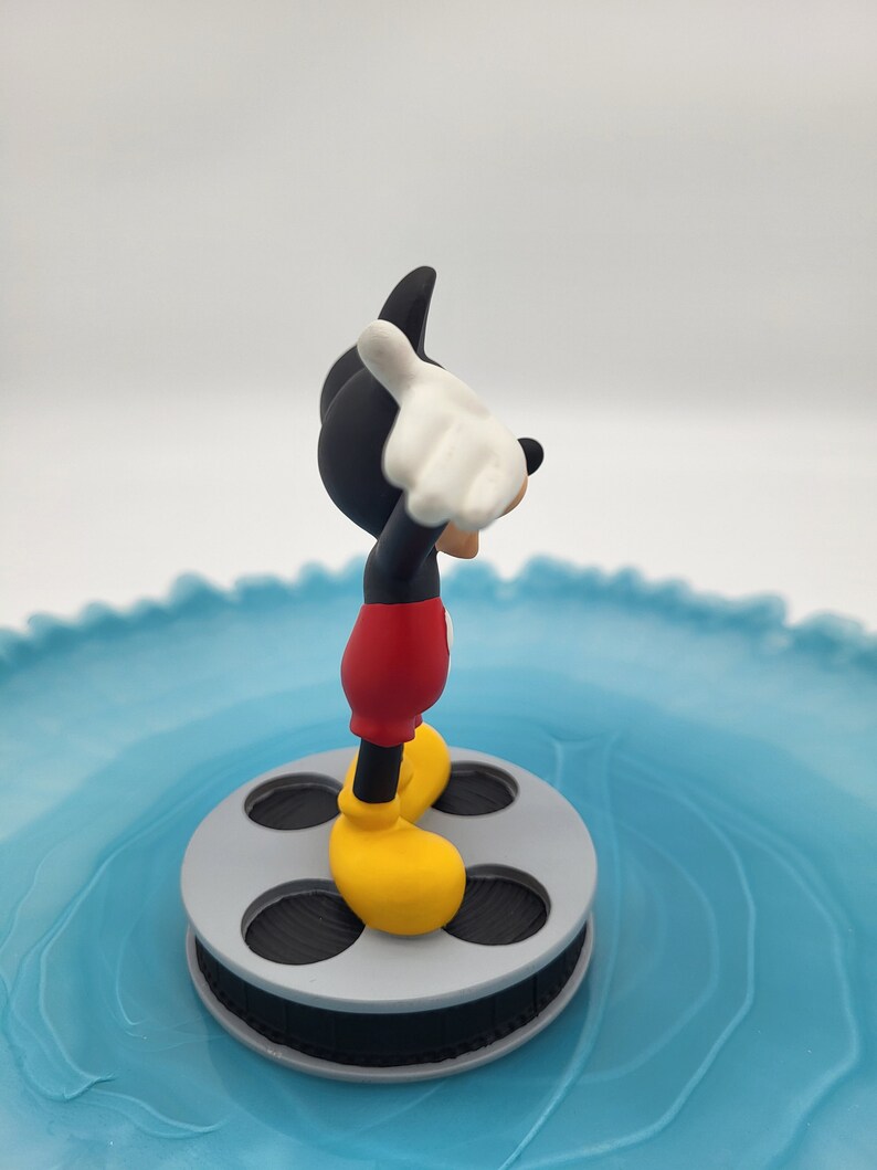 May include: A Mickey Mouse figurine, in his iconic outfit, stands on a grey film reel. Mickey is wearing red shorts, yellow shoes, white gloves, and a black body and head. The figurine is set against a light blue, swirled background.