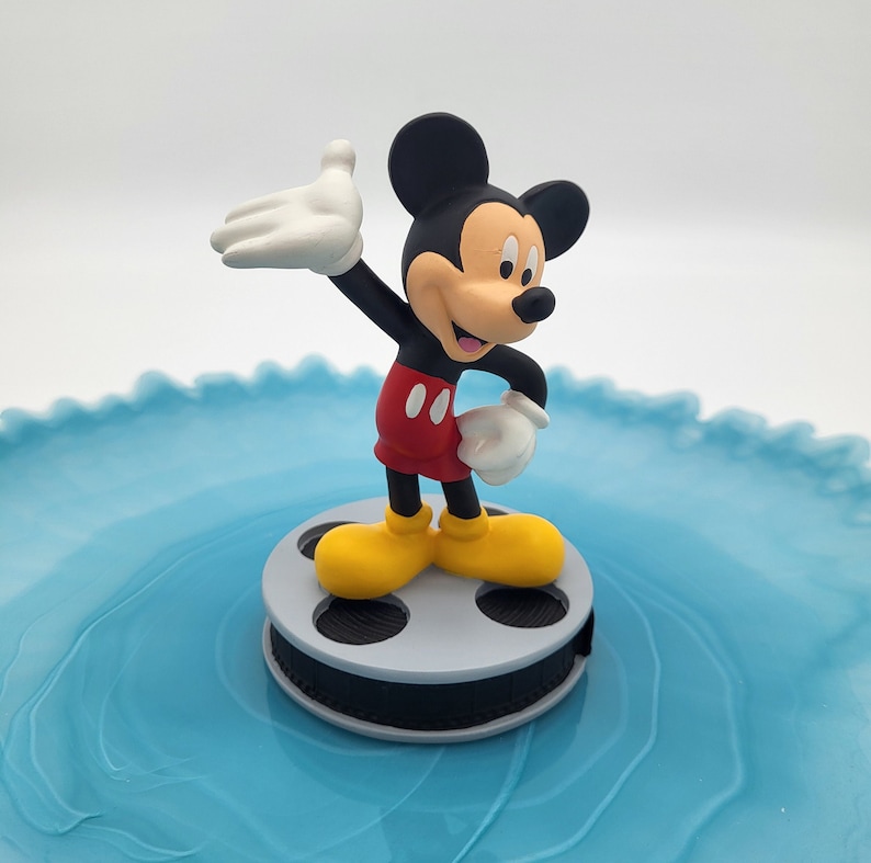 May include: A Mickey Mouse figure in a classic pose, wearing red shorts, yellow shoes, and white gloves. The figure stands on a grey film reel, set against a light blue, wavy background. The iconic character is a popular collectible.
