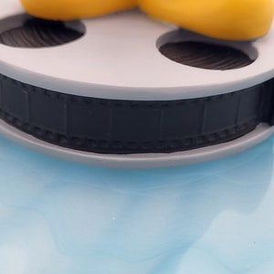 May include: A grey film reel with a black film strip. The reel is set against a light blue background. The film strip is detailed with individual frames. The reel is a decorative item.