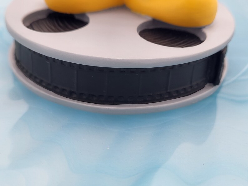 May include: A grey film reel with a black film strip. The reel is set against a light blue background. The film strip is detailed with individual frames. The reel is a decorative item.