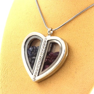 May include: Silver heart-shaped pendant necklace with a clear glass front, divided into two sections. One side contains dark blue stones, the other contains red stones. A line of small clear crystals separates the two sections. The necklace hangs on a silver chain.