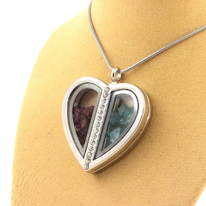 May include: A silver heart-shaped locket necklace with a clear glass front, divided into two sections. One side contains red gemstones, the other blue-green gemstones. A row of small clear stones decorates the center. The necklace hangs on a silver chain.