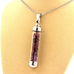 May include: A silver-toned necklace featuring a clear glass vial pendant filled with small, deep red gemstones. The vial is capped with silver metal and hangs from a delicate chain. The necklace is displayed on a beige mannequin.