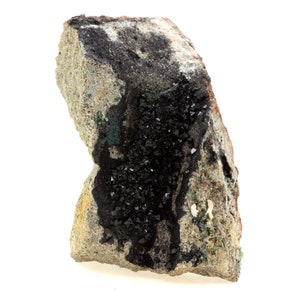 May include: A rough-hewn rock formation with a mix of colors, including gray, tan, and patches of green. The rock is speckled with small, dark, crystalline structures, creating a textured appearance. The overall shape is irregular and natural.
