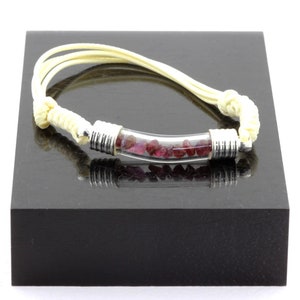 May include: A bracelet featuring a clear, curved glass tube filled with small, deep red gemstones. The tube is capped with silver-colored metal and connected to cream-colored cords. The bracelet is displayed on a black surface.