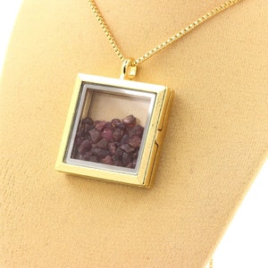 May include: A gold-colored necklace with a square pendant. The pendant is a clear box filled with small, dark red gemstones. The necklace chain is also gold-colored and has a delicate, box-link design. The pendant is displayed on a beige surface.