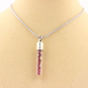 May include: A silver necklace with a clear glass pendant filled with small, pinkish-red gemstones. The pendant has a silver cap. The necklace is displayed on a neutral-colored background.