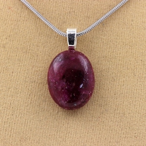Peut inclure: Un collier en argent avec un pendentif. Le pendentif est une pierre précieuse ovale, rouge foncé. La pierre est sertie dans une lunette en argent et suspendue à une chaîne en argent. Le fond est de couleur beige.