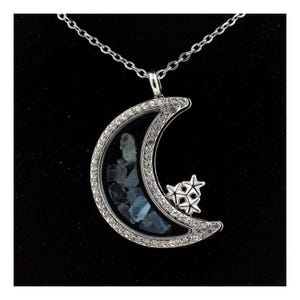 May include: A silver-toned necklace featuring a crescent moon pendant. The moon is encrusted with clear crystals and contains blue-grey stones. A cluster of stars is attached to the moon. The necklace hangs on a delicate chain.