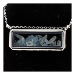 May include: A silver-toned necklace featuring a rectangular pendant filled with light blue, irregularly shaped stones. The pendant is encased in a black border and hangs from a delicate chain. The necklace is displayed against a black background.