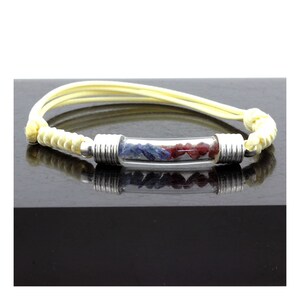 May include: A bracelet featuring a clear glass tube filled with blue and red gemstones. The tube is capped with silver-colored metal and attached to a yellow cord. The bracelet is displayed on a dark surface.