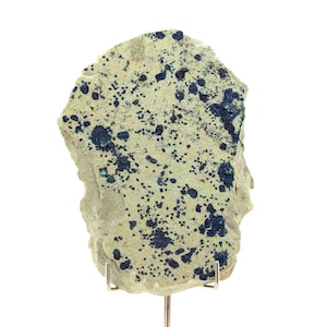 May include: A rough-cut mineral specimen, featuring a light green base color speckled with dark blue and green clusters. The rock is mounted on a silver stand, showcasing its natural, irregular shape. The mineral's surface has a textured appearance.