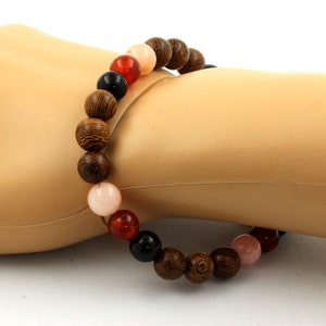 Peut inclure: Un bracelet de perles composé de perles en bois, rouges, noires et roses. Les perles en bois présentent un motif rayé. Le bracelet est présenté sur une surface claire.