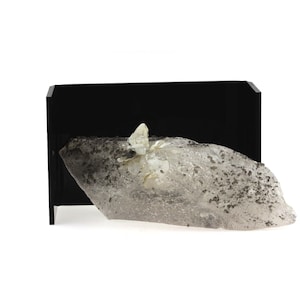 May include: A clear quartz crystal with a cluster of smaller crystals on top, encased in a black display box. The quartz is partially covered in a grey, speckled material.