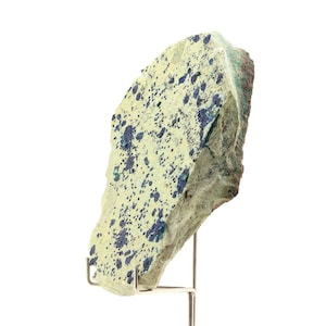 May include: A rough-edged rock sample with a light yellow-green surface speckled with dark blue and green mineral deposits. The rock is mounted on a silver metal stand, showcasing its natural texture and color variations. The specimen is ideal for collectors.