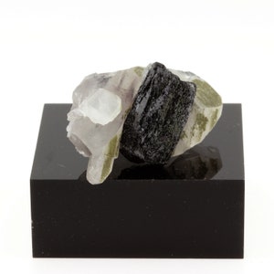 May include: A collection of raw gemstones, including a large black tourmaline crystal, is displayed on a glossy black square plinth. The gemstones also feature clear quartz and green-toned minerals. The background is plain white.