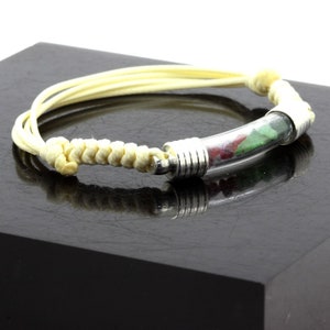 May include: A handmade bracelet featuring a clear glass tube filled with red and green stones, accented with silver-toned metal. The bracelet has a cream-colored woven cord and multiple cream-colored cords for the band.