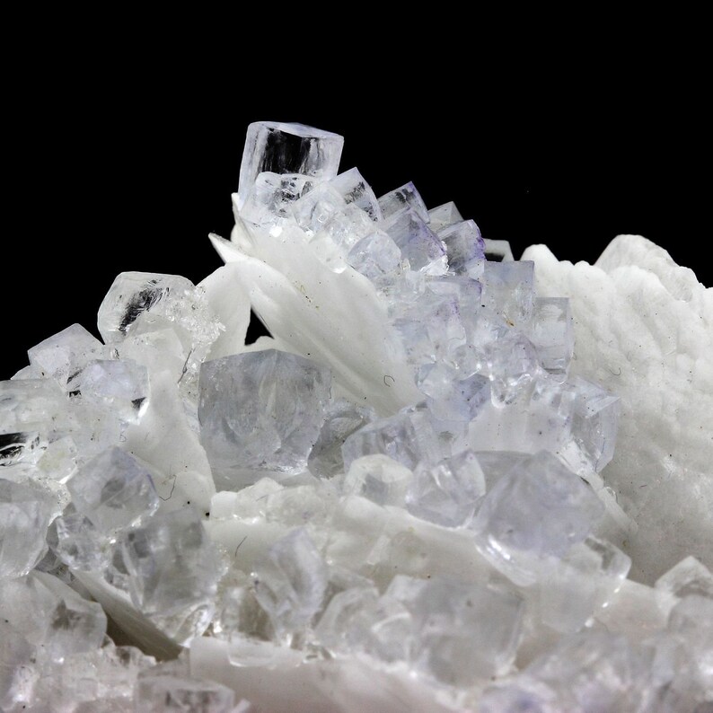 Puede incluir: Un grupo de cristales transparentes y transl&uacute;cidos con un tono ligeramente p&uacute;rpura. Los cristales est&aacute;n dispuestos en un patr&oacute;n aleatorio y se colocan sobre un fondo negro.