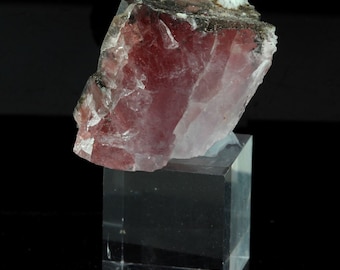 May include: A large, translucent pink crystal with a rough, uneven surface. The crystal is sitting on a clear acrylic display stand.