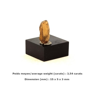 May include: A golden-brown gemstone, approximately 15 x 5 x 3 mm, is displayed on a black square base. The average weight of the gemstone is 3.54 carats. The gemstone has a rough, natural texture and is mounted on a small, light-colored support.